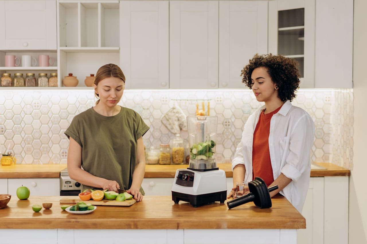 Two women preparing a healthy meal together in a modern kitchen, using fresh vegetables and a blender.